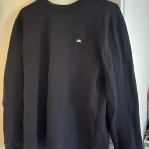 Svart sweatshirt från J.Lindeberg - Svart sweatshirt från J.Lindeberg med diskret logga på bröstet. Tröjan har rund hals och långa ärmar, tillverkad i mjuk bomull som är skön mot huden. Perfekt för dig som gillar stilrena och enkla plagg med en touch av sportig vibe.