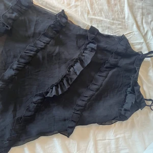 Svart volangklänning från Gina Tricot - Säljer en svart kortklänning från Gina Tricot med tunna axelband och flera diagonala volangdetaljer framtill. Klänningen har en lätt och luftig känsla, perfekt för dig som gillar en stilren look med extra detaljer. Materialet känns som polyester.