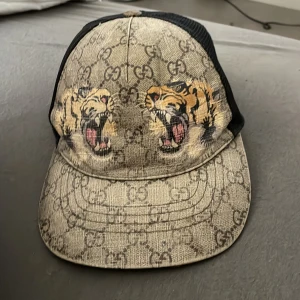 Gucci keps med tigertryck och GG-mönster äkta Gucci kep - Säljer en snygg Gucci keps med klassiskt GG-mönster i beige och brunt, prydd med två coola tigerhuvuden framtill. Baksidan har svart mesh och justerbart band med gröna och röda ränder. Perfekt statement piece för dig som gillar lyx och streetstyle.