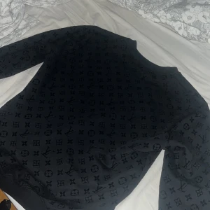 Svart Louis Vuitton sweatshirt - Svart sweatshirt från Louis Vuitton med ikoniskt monogrammönster över hela tröjan. Rund halsringning och långa ärmar. Mjuk och skön känsla, perfekt för dig som gillar lyxiga streetwear vibes. Passar dig som vill sticka ut med designerstil.