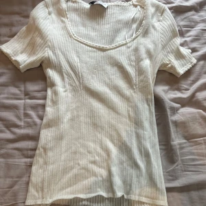 Vit ribbad topp med kort ärm - Säljer en ljusvit ribbad topp med korta ärmar och snygg fyrkantig ringning. Toppen har diskreta sömdetaljer framtill och är gjord i mjukt, stretchigt material som sitter skönt mot kroppen. Perfekt att styla med jeans eller kjol för en clean look.