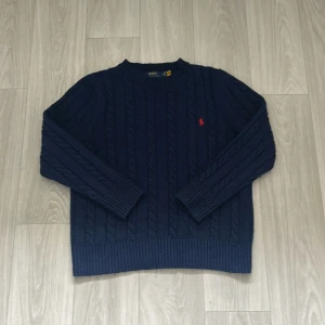 Mörkblå stickad tröja från Polo Ralph Lauren - Säljer en mörkblå kabelstickad tröja från Polo Ralph Lauren med klassisk röd broderad logga på bröstet. Tröjan har rund hals, långa ärmar och ribbade muddar. Perfekt för dig som gillar stilrena och tidlösa plagg med premiumkänsla.