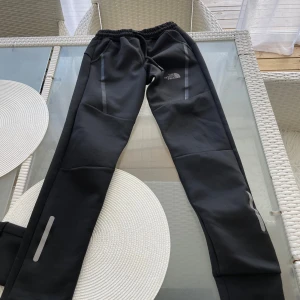 Två Svarta träningsbyxor från The North Face - Två Svarta träningsbyxor från The North Face med smal passform och elastisk midja. Byxorna har gråa reflexdetaljer längs benen och logga på höger lår. Perfekta för sport eller chill, tillverkade i mjukt syntetmaterial som andas.