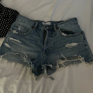 Blå slitna jeansshorts från Zara - Säljer ett par blå jeansshorts från Zara i storlek 38. De har hög midja, råa kanter och flera slitningar för en riktigt edgy vibe. Klassisk femficksmodell med dragkedja och knapp. Perfekta för sommaren och ger en avslappnad look.