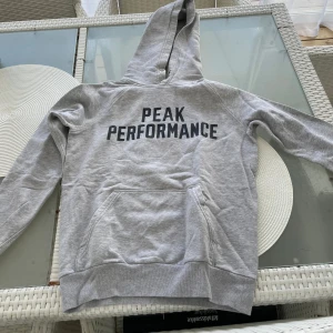 Grå hoodie från Peak Performance - Snygg grå hoodie från Peak Performance med stor svart logga på bröstet. Tröjan har huva med snörning och en klassisk magficka. Perfekt för chill eller träning, med mjukt bomullsmaterial och ribbade muddar vid ärmslut och nederkant.