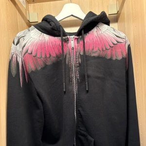Svart hoodie  - Säljer en svart hoodie från Marcelo Burlon med fett tryck av rosa och vita vingar över axlarna och ryggen. Tröjan har dragkedja framtill, huva med snörning och långa ärmar. Självklart äkta 