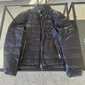 Svart pufferjacka från Prada - Säljer en marinblå pufferjacka från Prada med glansig finish och quiltad design. Jackan har ståkrage, två snedställda fickor med dragkedja och ikonisk Prada-logga på ärmen. Perfekt för dig som vill ha en clean och lyxig look under kyliga dagar. Defekter finns på bilderna, KVITTO FINNS !