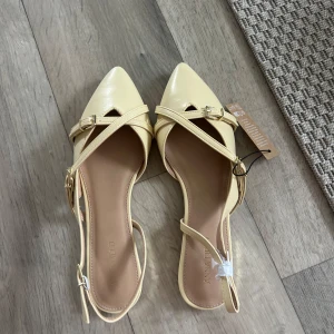 Gula ballerinaskor från Anna Field - Snygga ljusgula ballerinaskor från Anna Field med spetsig tå och smala remmar med spänne. Skorna är tillverkade i återvunnen polyester och har en platt sula. Perfekt för dig som gillar stilrena detaljer och vill ha något som sticker ut.