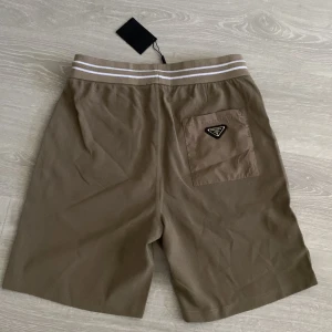 Prada shorts  - Snygga beige shorts med elastisk midja och vita ränder upptill. Shortsen har snörning framtill, sidofickor och en bakficka med klassisk Prada-logga. Tillverkade i ett mjukt material som känns lyxigt och bekvämt.