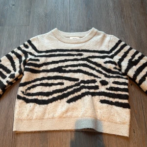 Beige zebramönstrad stickad tröja - Säljer en beige stickad tröja från Object i storlek S med svart zebramönster på framsidan och ärmarna. Tröjan har rund hals och ribbade muddar vid hals, ärmslut och nederkant. Perfekt för dig som gillar djurmönster och vill ha en chill look.