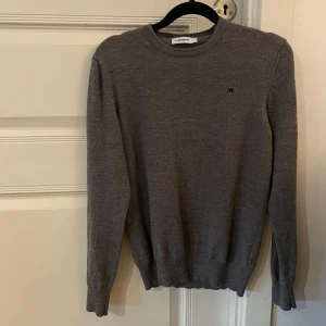 Grå stickad tröja från J.Lindeberg - Snygg grå stickad tröja från J.Lindeberg i storlek XS. Tröjan har rund hals, diskret logga på bröstet och ribbade muddar vid ärmslut och nederkant. Perfekt för dig som gillar stilrena och klassiska plagg med en modern touch. Tröjan passar Xs