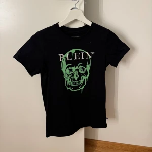 Svart t-shirt med grön döskalle Plein - Storlek 12år men passat mig som har XS. Cool svart t-shirt från Philipp Plein Junior med rund hals och korta ärmar. Framsidan har ett stort grönt döskalletryck och vit Plein-logga. 