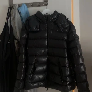 Svart bady Moncler  - Säljer en svart bady jacka från Moncler med glansig finish och klassisk logga på ärmen. Jackan har dragkedja framtill, praktiska fickor med dragkedja och en cool quiltad design. Perfekt för dig som vill ha en snygg och varm jacka med streetkänsla.