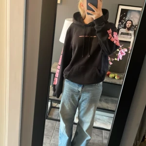 Svart hoodie med rosa tryck från H&M - Svart hoodie från H&M med stor rosa UNWIND-text på båda ärmarna och trycket 'FEEL THE PRESSURE DROP' på bröstet. Baksidan har ett stort blommigt motiv i svartvitt med rosa detaljer. Tröjan har huva med snörning och regular fit. Köpt på herravdelningen för en oversized look.