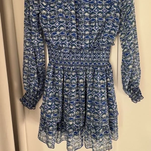 Blå mönstrad kortklänning från Zara - Säljer en blå och vit kortklänning från Zara med grafiskt mönster. Klänningen har långärmade ärmar med smock vid handlederna, markerad midja och v-ringad hals. Materialet är lätt och luftigt, perfekt för vår och sommar. Supersnygg till sneakers eller boots!