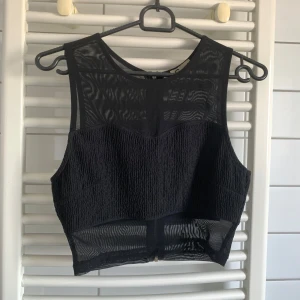 Svart croppad mesh topp med dragkedja - Snygg svart croppad topp med transparent mesh upptill och nedtill. Toppen har en cool struktur på mittenpartiet och en dragkedja i ryggen. Ärmlös design som ger en edgy vibe, perfekt att styla med höga jeans eller kjol.