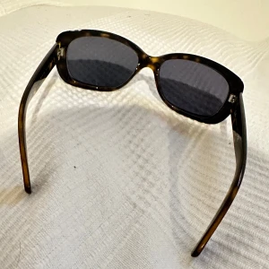 Bruna Ray-Ban solglasögon - Snygga solglasögon från Ray-Ban med brunmelerad båge i plast och svarta glas. Modellen har en mjukt rektangulär form som ger en cool vintagekänsla. Perfekt accessoar för att höja din stil på soliga dagar.