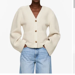 Beige stickad kofta med v-ringning - Säljer en beige stickad kofta med v-ringning och stora träknappar framtill. Koftan har två fickor och breda, långa ärmar som ger en chill och modern vibe. Perfekt att slänga på över en t-shirt eller linne för en enkel och snygg look.