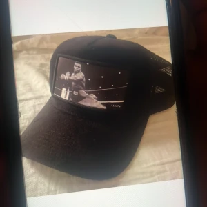 Svart keps med boxningsmotiv - Cool svart keps med mesh på sidorna och ett stort boxningsmotiv framtill. Perfekt för dig som gillar sportiga accessoarer och vill sticka ut med en unik design. Märket ALG74 syns på framsidan av kepsen.