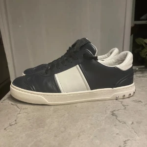 Valentino Garavani sneakers blå/vit - Snygga Valentino Garavani sneakers i mörkblått och vitt skinn med coola nitar på sulan och camouflagemönstrad undersida. Klassisk låg modell med rund tå och svarta snören. Perfekt för dig som gillar exklusiva detaljer och streetstyle.