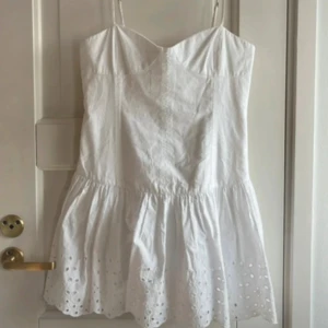 Vit kort klänning med broderi - Säljer en söt vit kort klänning med tunna axelband och broderade detaljer längst ner på kjolen. Klänningen har en luftig passform och är tillverkad i bomull, perfekt för varma dagar. .
