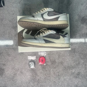 Nike Jordan 1 Low Travis Scott - Nike Jordan 1 Low Travis Scott med omvänd vit Swoosh, grå mocka och vita skinnpaneler. Röda detaljer på sidan och tungan, samt extra skosnören i svart och rött medföljer. Platt sula och rund tå, perfekt för dig som vill sticka ut med en ikonisk sneaker.