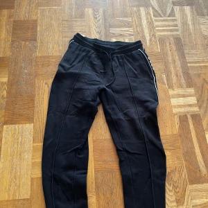 Svarta mjukisbyxor från BOSS - Svarta mjukisbyxor från BOSS med vita logotexter längs sidorna och snygga ribbade muddar i midjan. Byxorna har snörning i midjan och är tillverkade i mjukt bomullsmaterial. Passformen är normal och de har en sportig vibe med raka ben och diskreta sömmar.