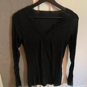 Svart långärmad topp med spets - Svart långärmad topp med v-ringning och snygga spetsdetaljer både vid halsen och ärmsluten. Tunn och mjuk, perfekt för att lyfta din outfit med en elegant touch. Sitter snyggt och har en feminin vibe.