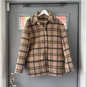 Rutig beige ulljacka från & Other Stories - Snygg rutig jacka från & Other Stories i beige och mörkgrå toner, tillverkad i mjuk ull från Manteco. Jackan har klassisk krage, knäppning med knappar och två stora fickor framtill. Perfekt för dig som gillar stilrena och tidlösa plagg.