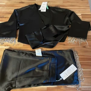 Svart satinkostym med pärldetaljer - Snygg svart kostym från NA-KD i glansigt satinmaterial. Kavajen har omlottmodell med breda slag och pärlfransar vid ärmsluten. Byxorna är raka och har matchande pärlfrans längs bensluten. Perfekt för dig som vill sticka ut med coola detaljer.