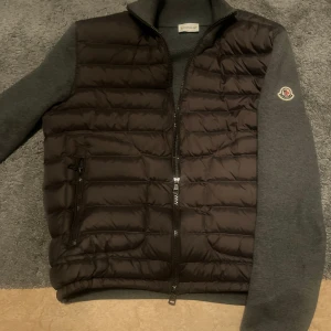 Moncler cardigan - Snygg grå cardigan från Moncler med quiltad framsida och dragkedja. Västen har en stilren design med ribbad krage och grå stickade partier på ryggen och ärmarna. Moncler-logga på vänster ärm ger extra premiumkänsla. Perfekt för lager-på-lager och streetstyle.