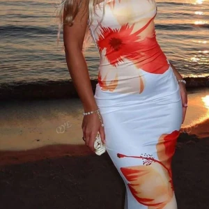 Vit långklänning med röda blommor - Supersnygg långklänning i vit med stora röda och orange blommor. Klänningen har en figurnära passform och är ärmlös, perfekt för varma dagar. Materialet känns mjukt och stretchigt, vilket gör den bekväm att bära. Perfekt för dig som vill sticka ut med färg och mönster.