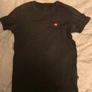 Svart t-shirt från Kronstad  - Svart t-shirt från Kronstad Kilo med rund hals och korta ärmar. På bröstet finns en liten röd patch med vitt tryck. T-shirten är gjord i mjuk bomull och har en clean, enkel look som funkar till det mesta. Passformen är normal och materialet känns skönt mot huden.