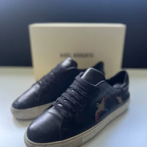 Svarta sneakers från Axel Arigato - Snygga svarta sneakers från Axel Arigato med broderade fåglar i rött, blått och beige på sidan. Skorna har snörning, rund tå och vit platt sula. Tillverkade i skinn för en lyxig känsla och stilren look.