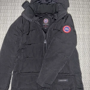 Svart dunjacka från Canada Goose - Säljer en svart dunjacka från Canada Goose med huva och klassisk logga på ärmen. Jackan har dragkedja framtill, två stora fickor med lock och en stilren, puffad design som håller dig varm. Perfekt för kalla dagar och snygg till streetwear.