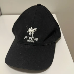 Svart keps Polo Club Vinson - Svart keps från Polo Club Vinson med vit broderad logga och polospelare framtill. Justerbar rem baktill med metallspänne och amerikansk flagga broderad ovanför öppningen. Klassisk böjd skärm och stilren look, perfekt för dig som gillar sportig stil.
