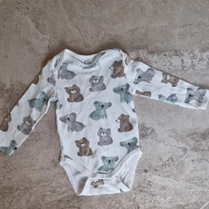 Vit långärmad body med björnar C&A - Vit långärmad body från C&A med gulligt mönster av grå, bruna och blå björnar och koalor.Bodyn har praktiska tryckknappar i grenen och är tillverkad i mjuk bomull som är skön mot huden. Bodyn är helt ny utan prislapp. 