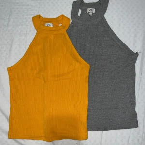 Ribbade ärmlösa toppar från River Island - Två snygga ribbade ärmlösa toppar från River Island, en i grått och en i starkt gul färg. Båda har hög hals och croppad passform, perfekta till jeans eller kjol. Materialet är mjukt och stretchigt, vilket gör dem riktigt bekväma. Säljer dem ihop