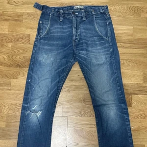 Blå jeans från Acne Jeans - Säljer ett par klassiska blå jeans från Acne Jeans med snyggt slitna detaljer och raka ben. Jeansen har knäppning med metallknappar, fem fickor och tydliga kontrastsömmar. Perfekta för dig som gillar en avslappnad och tidlös look.