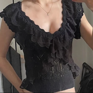 Svart spetsig blus med volang - Supersöt svart blus med bred volang runt v-ringad hals och axlar. Spetsdetaljer och ribbad nederkant ger en cool vintagekänsla. Ärmlös och croppad modell.