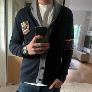 Stickad kofta - Snygg cardigan från GANT i marinblått med grå krage och knappslå. Långärmad med grå och röda ränder på ena ärmen samt ett broderat GANT-emblem på bröstet. Tillverkad i mjuk bomull, perfekt för dig som gillar klassisk college-stil. Perfekt för hösten och vintern