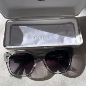 Grå transparenta solglasögon CHIMI - Snygga solglasögon från CHIMI med transparent grå båge och mörka, lätt tonade linser. Modellen har en modern, fyrkantig form och levereras i originalfodral med putsduk. Perfekt accessoar för att lyfta din stil i solen.