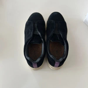 Svart mocka sko herr. -  Hej! Säljer ett par svarta sneakers från Eytys i mocka med vit platt sula och en lila rem baktill. Skorna har en minimalistisk design utan snörning och insida i skinn. Paketstorlek: längd=32cm bredd=25cm höjd=14cm!