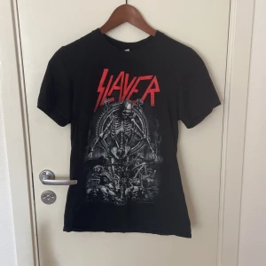 Svart Slayer t-shirt från Gildan - Cool svart Slayer t-shirt från Gildan med stort rött Slayer-tryck och mörkt skelettmotiv både fram och bak. Klassisk rund hals och korta ärmar. Tillverkad i mjuk bomull, perfekt för dig som gillar metal och vill sticka ut med en fet bandtröja.
