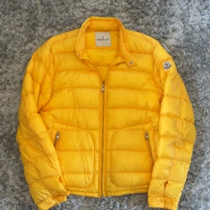 Gul Moncler dunjacka i storlek L - Säljer en riktigt snygg gul dunjacka från Moncler. Jackan har quiltad design, dragkedja framtill och två fickor med dragkedja. Klassisk Moncler-logga på ärmen och hög krage med tryckknapp. Perfekt för dig som vill sticka ut i vinter!