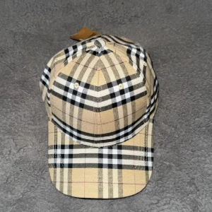 Beige rutig Burberry keps - Snygg keps från Burberry i klassiskt rutigt mönster med beige, svart och vit färg. Kepsen har böjd skärm och justerbar rem baktill. Tillverkad i bomull med typisk Burberry-look som ger en trendig vibe till din outfit. 100 procent äkta köpt på NKs för några tusen. Säljer 500kr då jag inte använder den