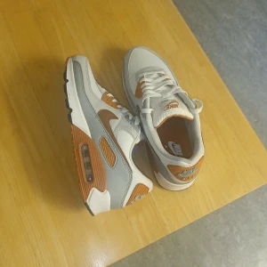 Nike Air Max 90 i grå och brun - Nike Air Max 90 sneakers med snygg mix av ljusgrått och brunt. Skorna har klassisk Air Max-sula med synlig luftbubbla, vita snören och detaljer i mocka och syntet. Perfekt för dig som gillar retrovibe och streetstyle. Swoosh-logga på sidan och Air Max-märke på hälen.
