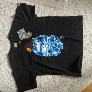 Svart BAPE t-shirt med blå camo print - Svart t-shirt från BAPE med ikonisk apa-grafik på bröstet och blått camouflage-mönster. Rund hals och korta ärmar. Märkets taggar och detaljer syns tydligt. Perfekt för dig som gillar streetwear och vill sticka ut med en fet design.