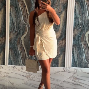 Ljus omlottklänning med oneshoulder - Säljer en superfin ljus kortklänning med omlott-design och oneshoulder. Klänningen har en snygg knyt-detalj i midjan och är tillverkad i ett mjukt, lätt material som ger en skön känsla. Perfekt för dig som gillar stilrena och trendiga plagg.