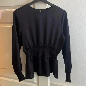 Svart blus med smockad midja - Säljer en svart blus med rund hals och långärmade ärmar. Blusen har smockad midja som ger en snygg siluett och volang vid nederkanten. Materialet är lätt och har en skön känsla mot huden. Perfekt för dig som gillar stilrena och enkla plagg.